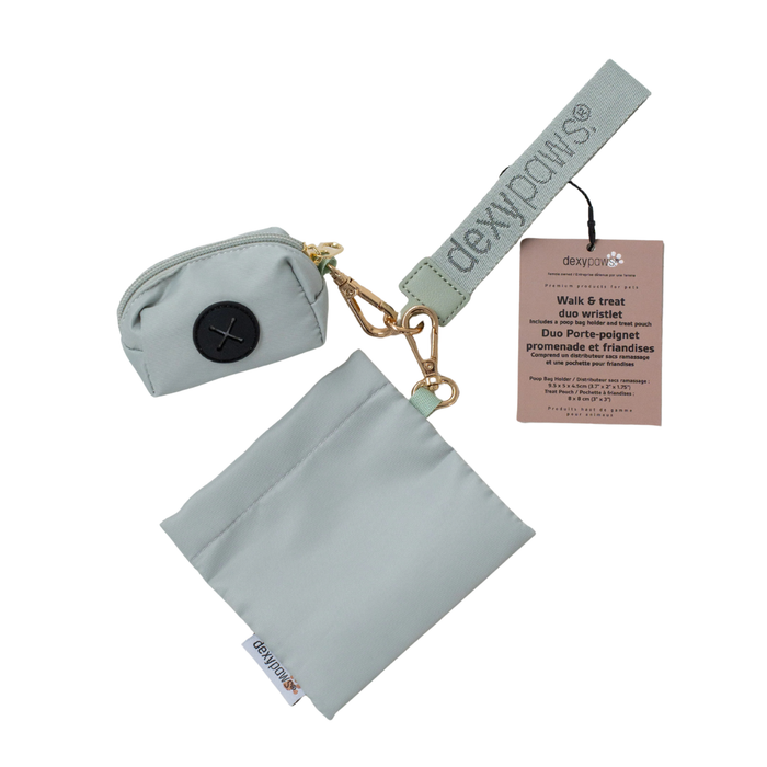 The product is a soft sage green in colour with brass coloured metal clips. It shows a wrist strap with "dexypaws" stitched into it with slightly darker sage thread. A poo bag holder and a pouch are attached on separate clips. The pouch is a flat square shape and has a Dexypaws branded tag stitched into the side. The poo bag holder has a sage-coloured zipper at the top, with a brass tab. The dispensing part is a black circle with an X shaped hole in the middle.